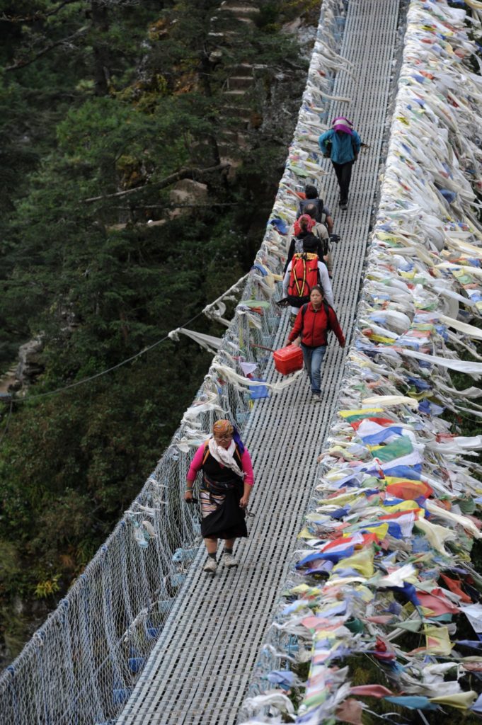 Attraversamento di un ponte sospeso in acciaio, con moltissime bandiere di preghiera, da parte di trekkers e portatori nel percorso di trekking da Phakding e Namche Bazar
