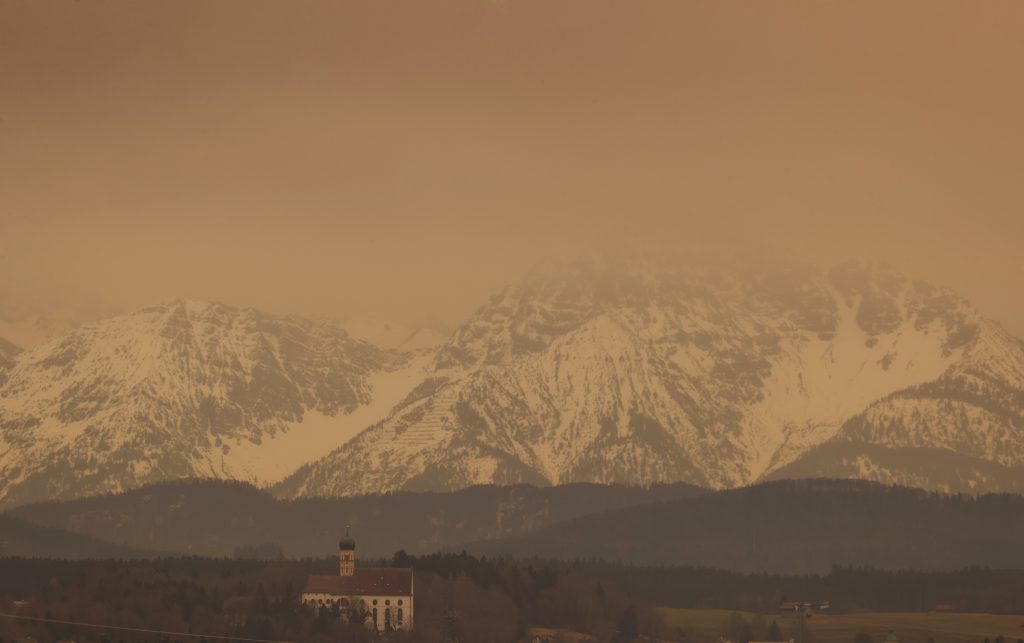 15 March 2022, Bavaria, Ruderatshofen: Dust from the Sahara colors the sky above the Alps and the parish church of St. Martin in Marktoberdorf in reddish hues. The associated clouding of the sky can make the sun appear milky even in otherwise cloud-free skies. Photo: Karl-Josef Hildenbrand/dpa