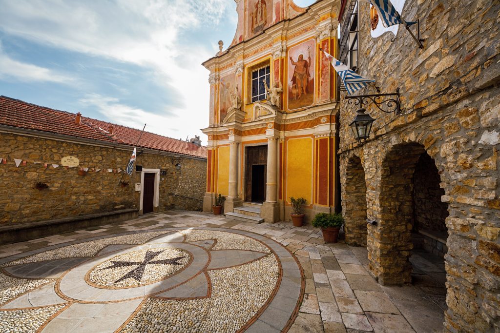 Costruita nel 1615, la chiesa di San Martino a Seborga ha sul prospetto dipinti del 1928 (Cristo redentore e Arcangelo Michele). Sulla piazza ovale, con un pavimento a mosaico, affaccia anche il palazzo dei Monaci, che fu sede della zecca dell’antico principato. Oggi è di proprietà privata, ma sventolano ancora le bandiere a strisce bianche e blu (www.principato diseborga.com). Foto Gabriele Bignoli/Adobe Stock