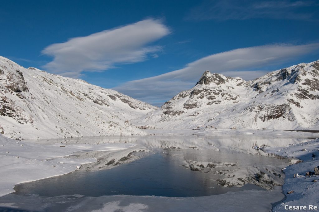 Il Lago dello Spluga, all’omonimo passo.  Tre situazioni diverse per tre paesaggi di lago. Nella prima foto la superficie è ghiacciata, ma non bianca di neve. Queste situazioni si verificano, in genere, a inizio stagione, con l’abbassarsi della temperatura e ancora poca neve al suolo. Il secondo lago è, invece, totalmente imbiancato, con la superficie liscia e bianca. La terza foto mostra, invece, una situazione mista con parti dello specchio d’acqua ghiacciate e parti innevate, con riflessi solo parziali.  