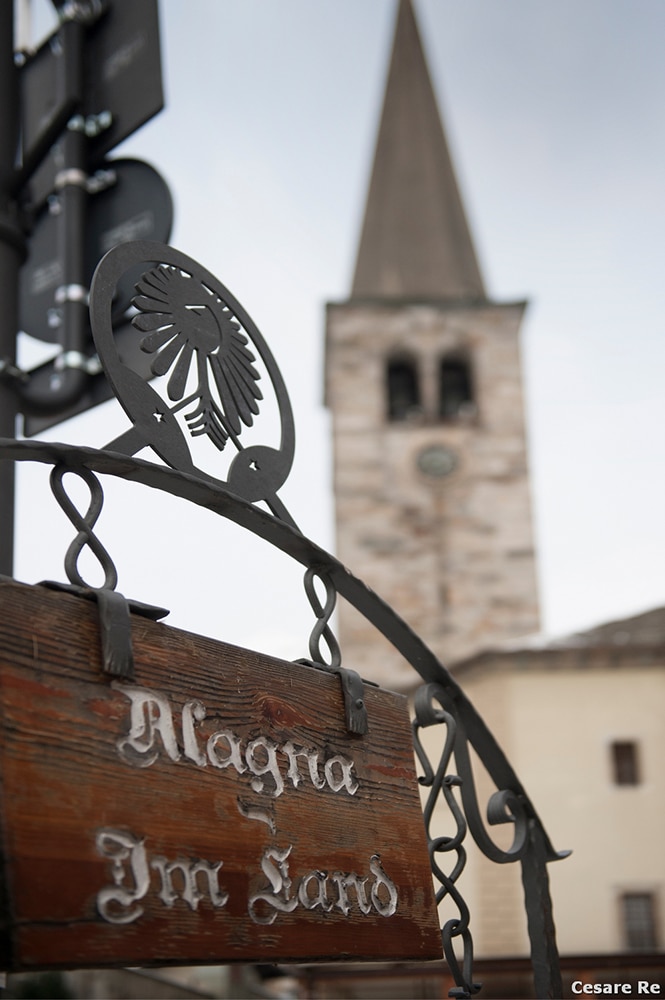 Im land. Il campanile di Alagna Valsesia. Il punto di messa a fuoco è sul cartello, con la scritta in lingua Walser di “Im land” (terra), nome col quale veniva chiamata questa località. Lo scatto sottolinea la presenza intensa e ancora attuale della cultura Walser, nel territorio della Valsesia. Il Campanile è ben riconoscibile e qualsiasi osservatore è consapevole del soggetto che si vede sullo sfondo. Nikon D800; Nikkor 24-70 2,8 AFG; foclae di 50 mm; 1/1250 sec; f/2,8; ISO 400.